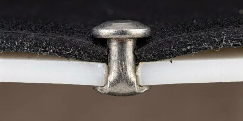 .Cross-section macro photo showing a metal rivet anchored through leather and into a rigid white PE board backing.