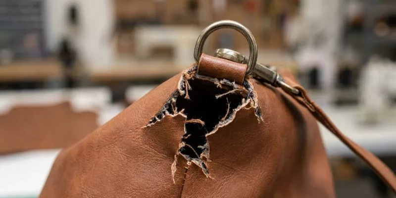 Failed bag attachment showing a metal D-ring tearing completely through a single layer of thin brown PU leather.