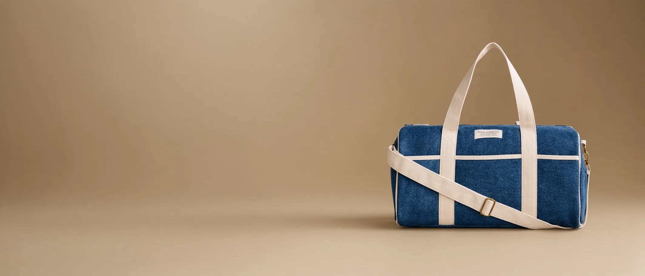 A finished duffel-style bowling bag in blue denim with cream webbing handles and a shoulder strap, on a warm tan studio ground