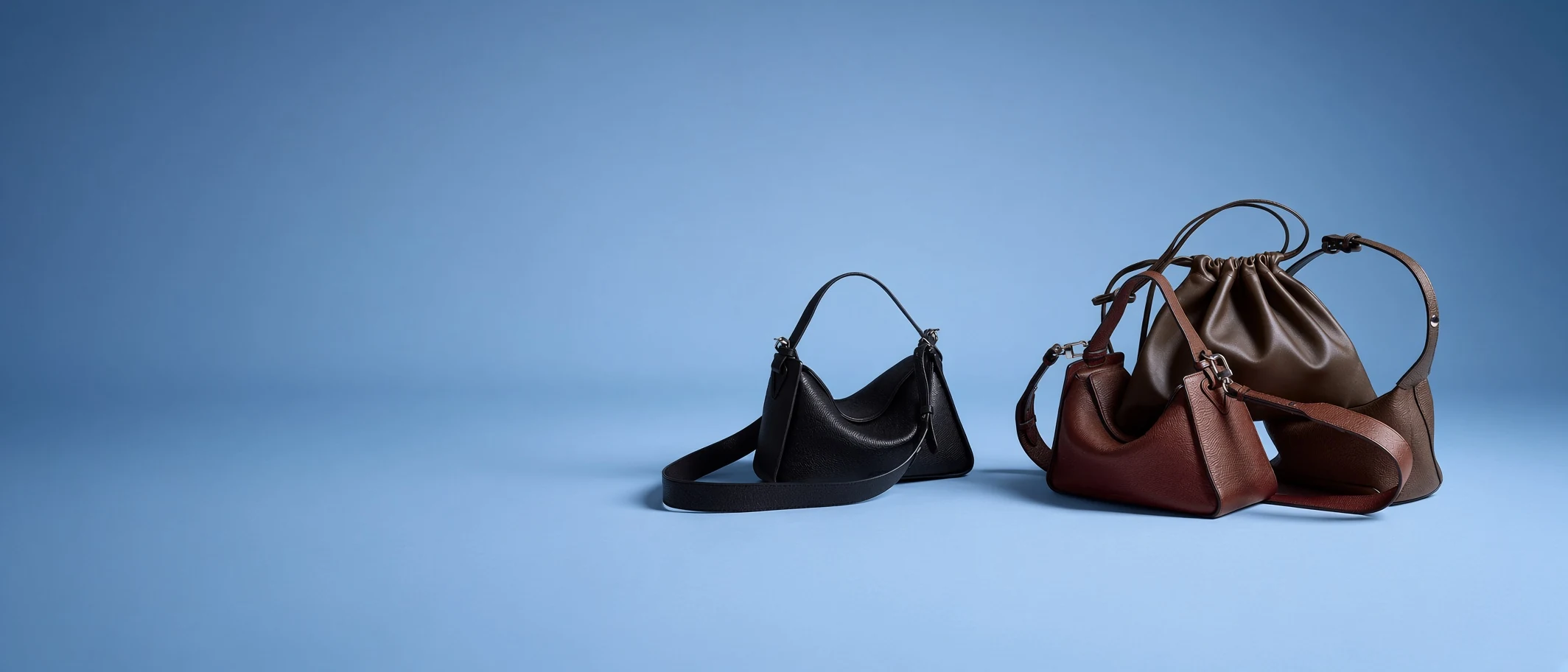 A group of leather crossbody bags on a pale blue ground — a black shoulder bag with a wide adjustable strap, a tan and a burgundy crossbody bag with buckled straps, and a brown drawstring pouch behind them