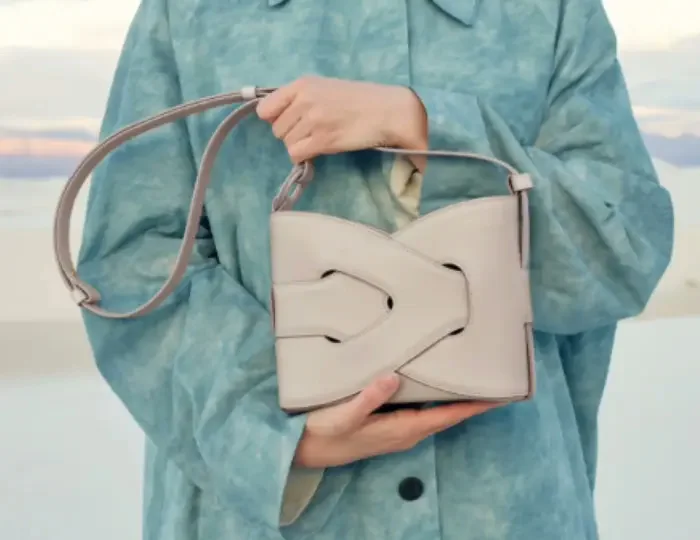 A person in a pale blue jacket holding a small structured crossbody bag by its strap against a soft background
