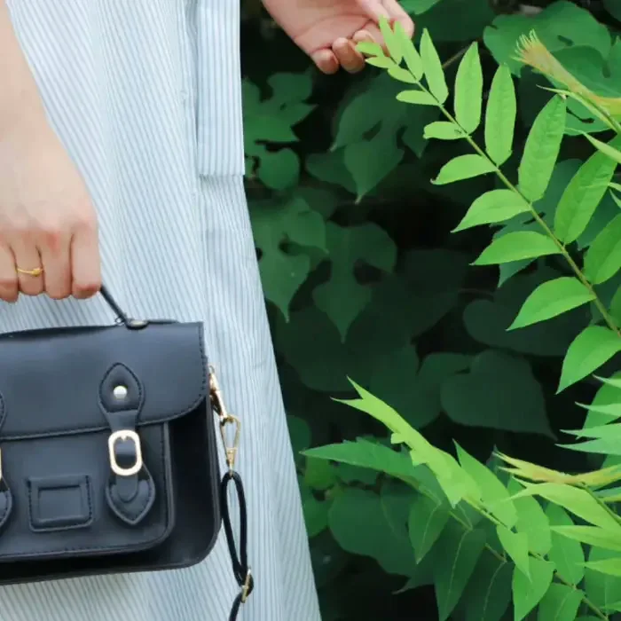 A hand lifting a black leather crossbody bag by its strap beside a wall of green leaves
