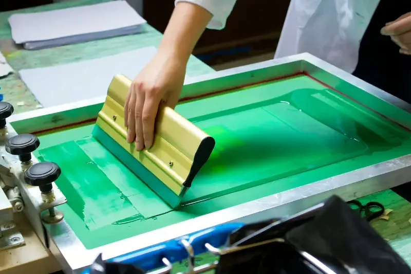 A printer pulling a squeegee across a green screen-printing frame to apply a logo to cloth