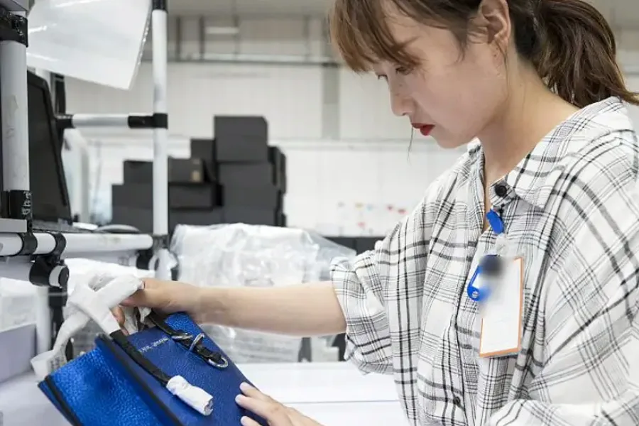 An inspector examining the stitching and hardware of a finished bag at a backlit quality-control bench