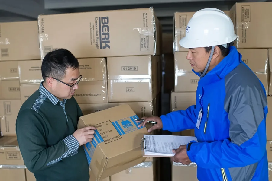 Two warehouse staff checking a carton against a printed checklist before loading, in front of stacked export cartons
