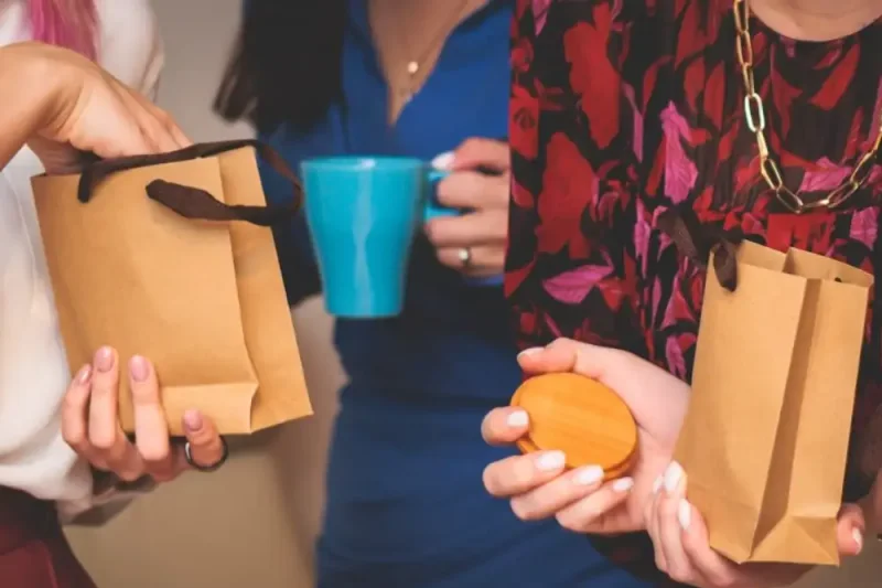 Several pairs of hands holding kraft gift bags and a blue travel cup at a corporate gifting hand-out