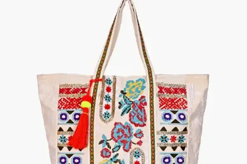 A cream canvas tote printed with an embroidered folk-floral pattern, held by its handles on a white background