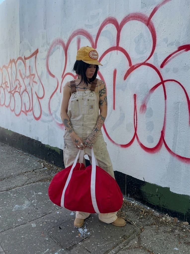 A red canvas duffle held by its rope handles against a graffitied wall
