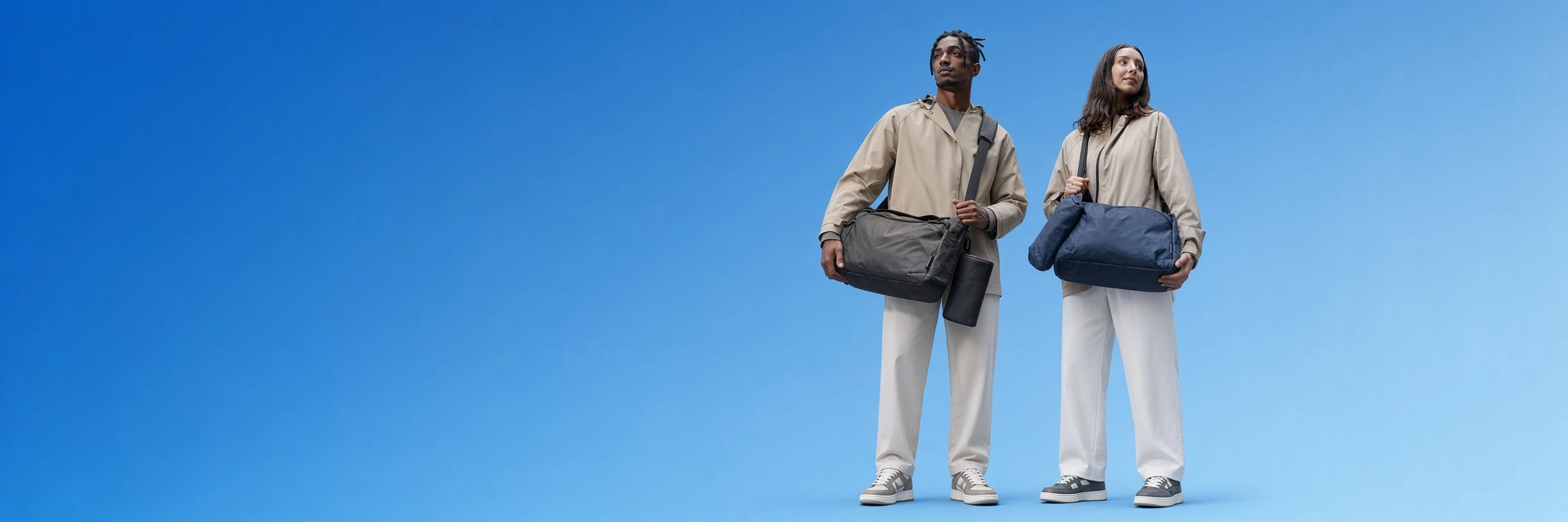 Two people in light jackets each holding a duffle bag, photographed against a blue studio backdrop