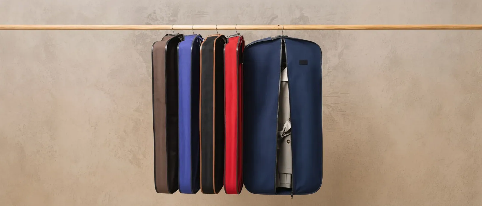 A rail of garment covers in brown, black, blue, red and navy, hung from a wooden rail against a bare plaster wall