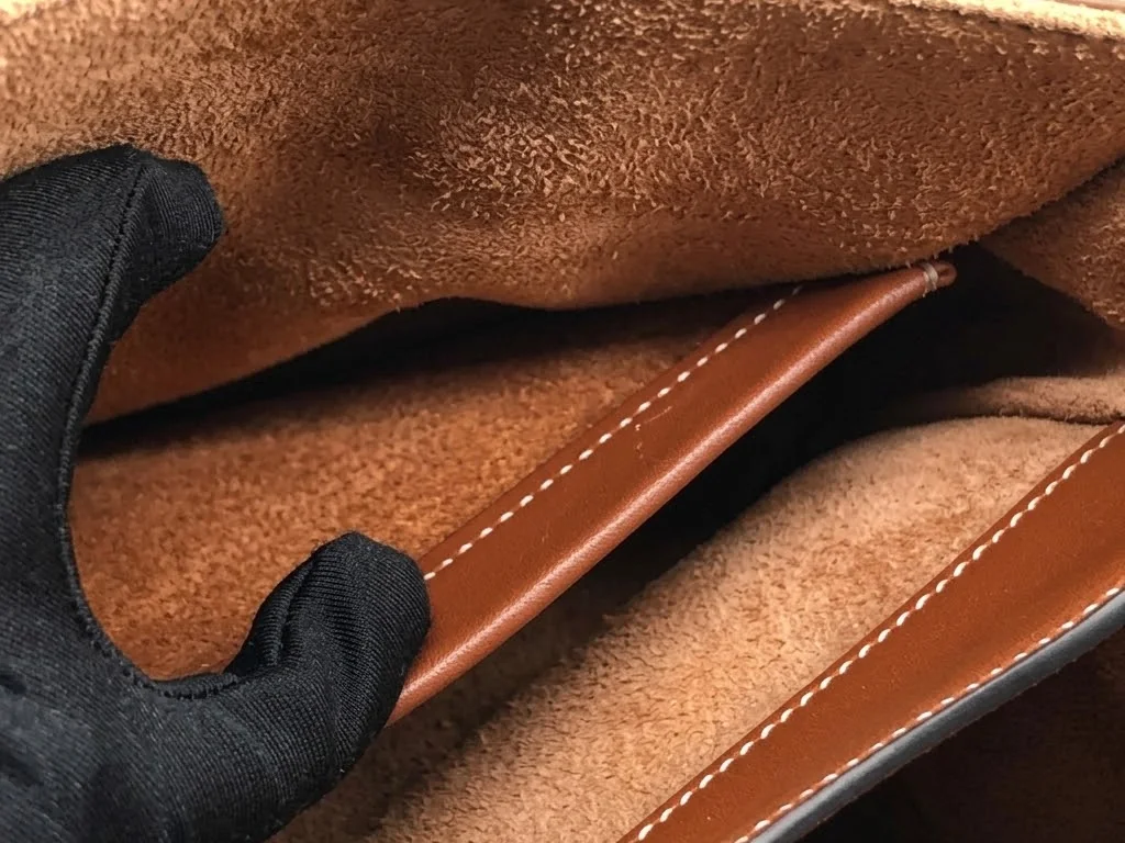 A gloved hand lifting the inner flap of a leather handbag to show its suede lining and stitched edge trim