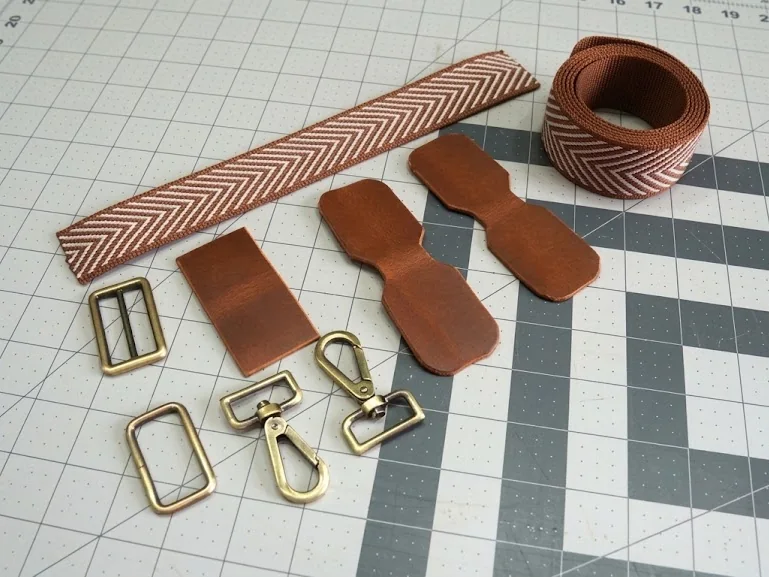 A woven webbing strap, leather strap tabs and antique brass buckles and snap hooks laid out on a cutting mat