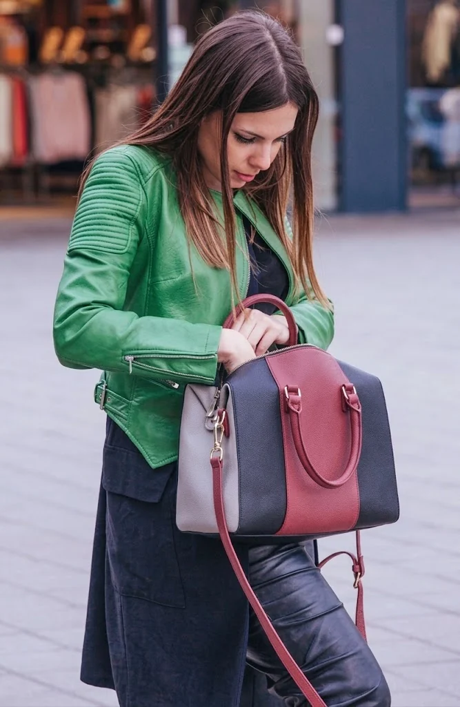 A three-panel leather handbag in grey, burgundy and black, held by its top handle on a city street