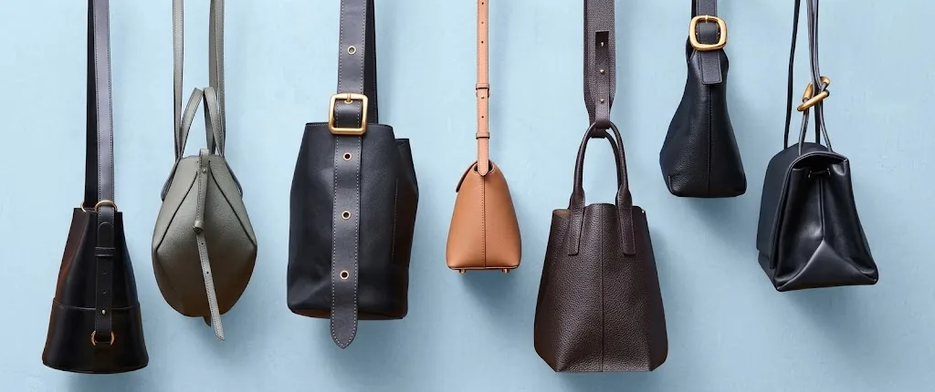 Seven custom leather handbags in tan, olive, black and brown hanging from their straps against a pale blue wall