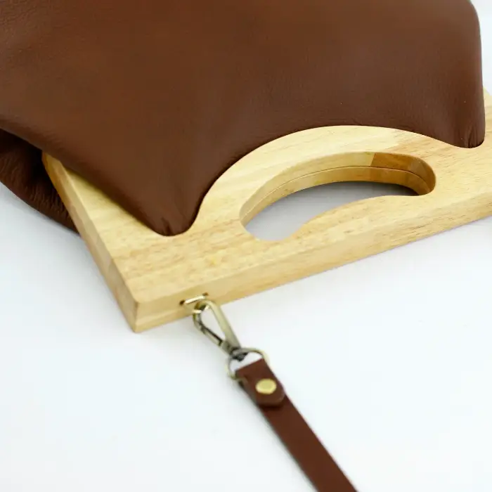 A brown leather bag resting on a light wooden frame with a cut-out handle, a swivel clip and strap beside it