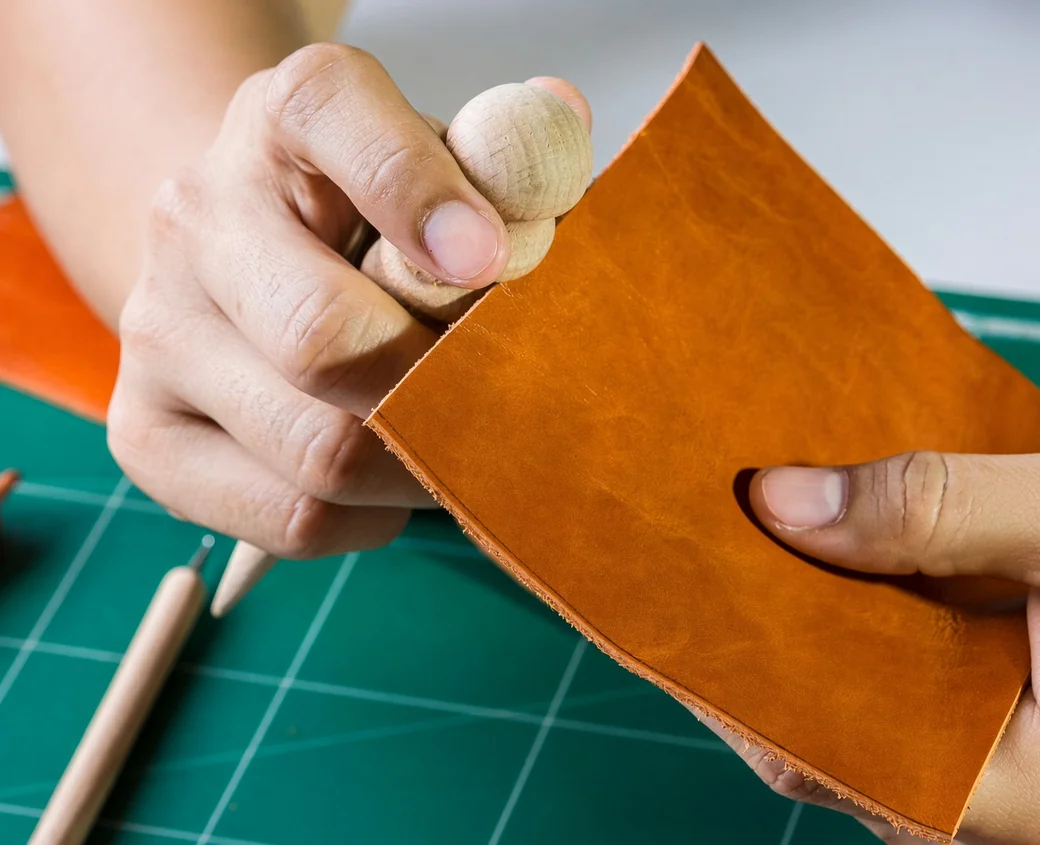 Hands holding a square of tan vegetable-tanned leather with its cut edge showing the thickness, over a green cutting mat with leather tools beside it