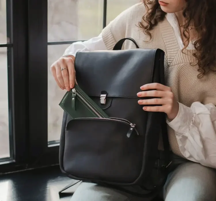 A person opening the front pocket of a black leather backpack to reach a small pouch inside