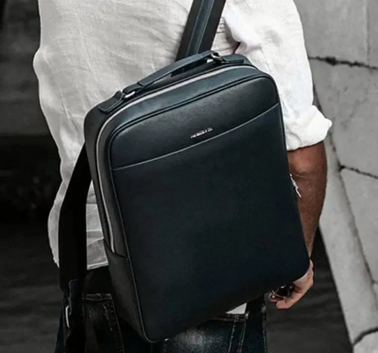 A person carrying a black leather laptop backpack over a white shirt and dark jeans