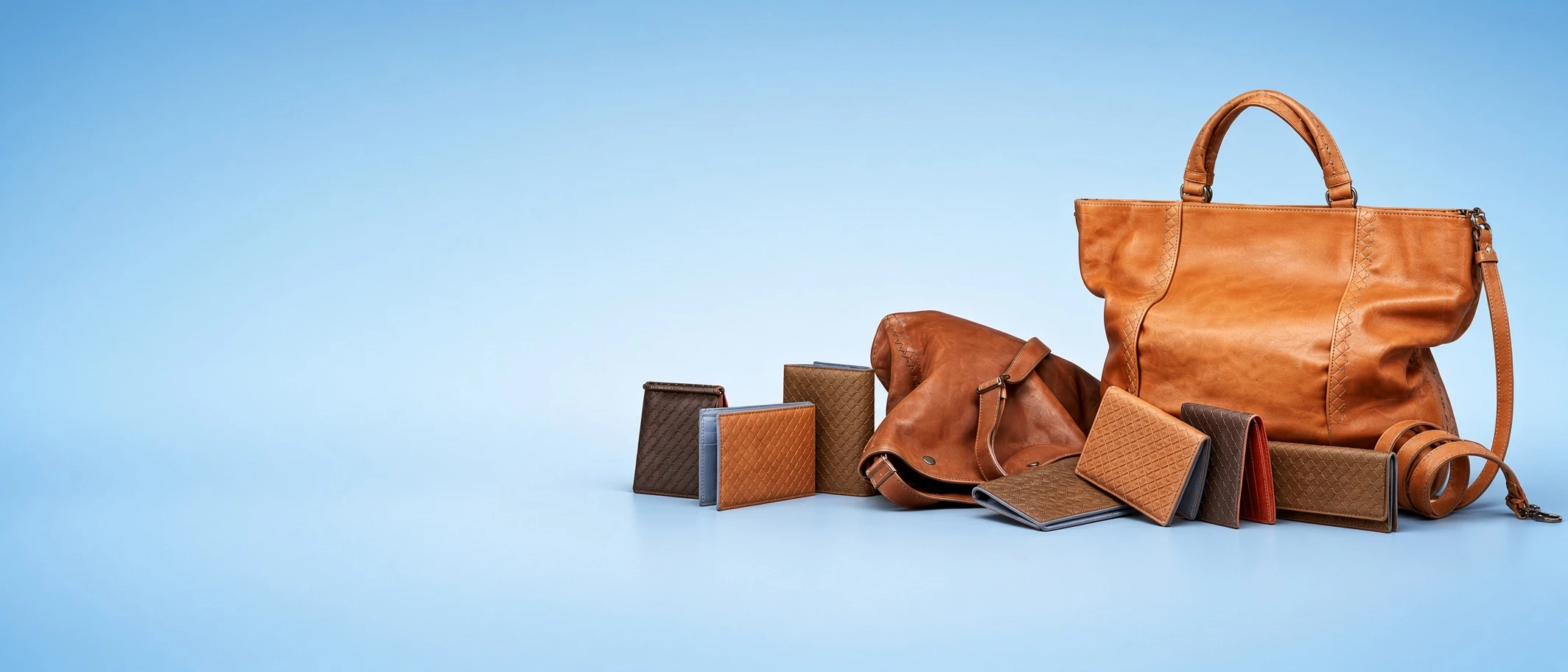 A group of leather goods — a tall tote, a messenger bag, wallets and belts — arranged on a pale blue studio sweep