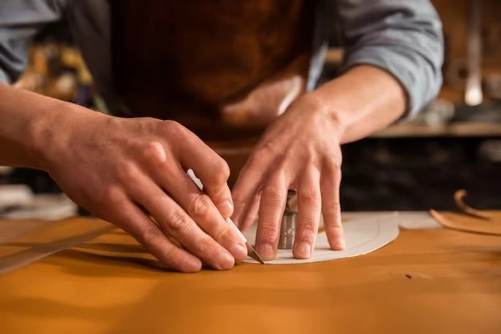 Two hands cutting a leather panel along a paper pattern with a blade on a workbench