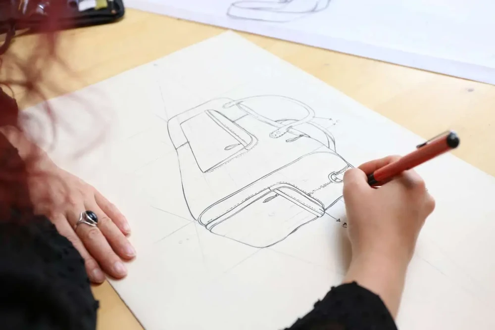 A designer drawing a bag on white paper with a red pen, a sketch of the finished shape above