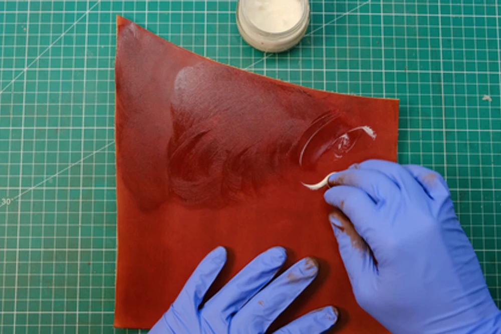 Gloved hands burnishing the cut edge of a red leather panel with a small tool