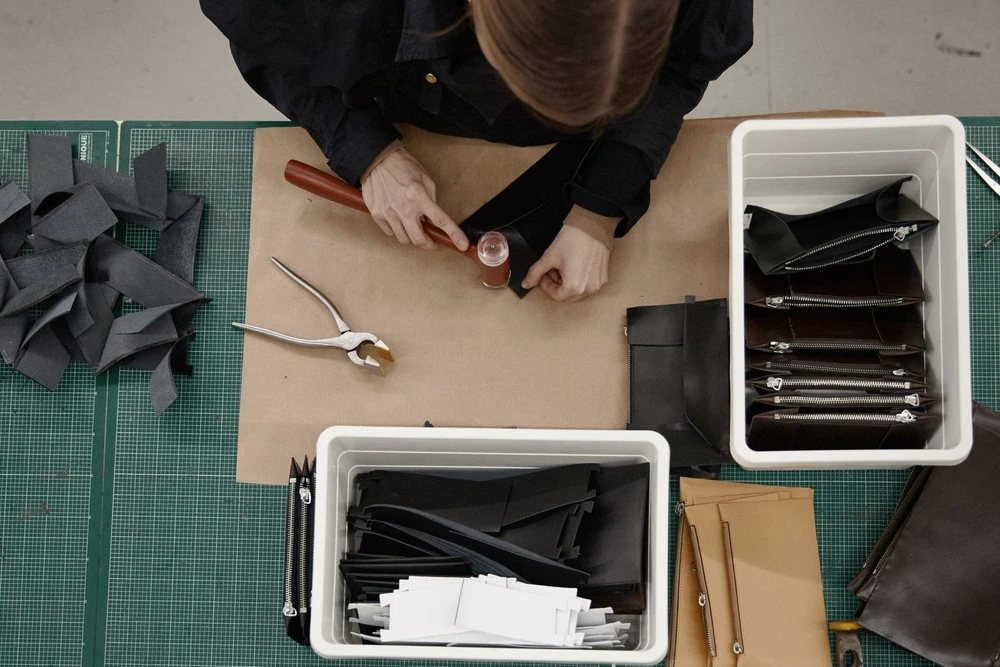 A workbench seen from above with leather panels, a hammer-set zip pull and finished pieces in trays