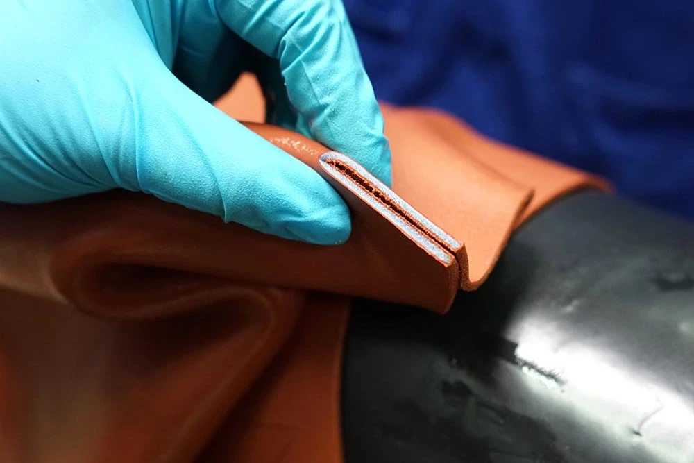 A gloved hand inspecting the cut edge of a tan leather panel against light