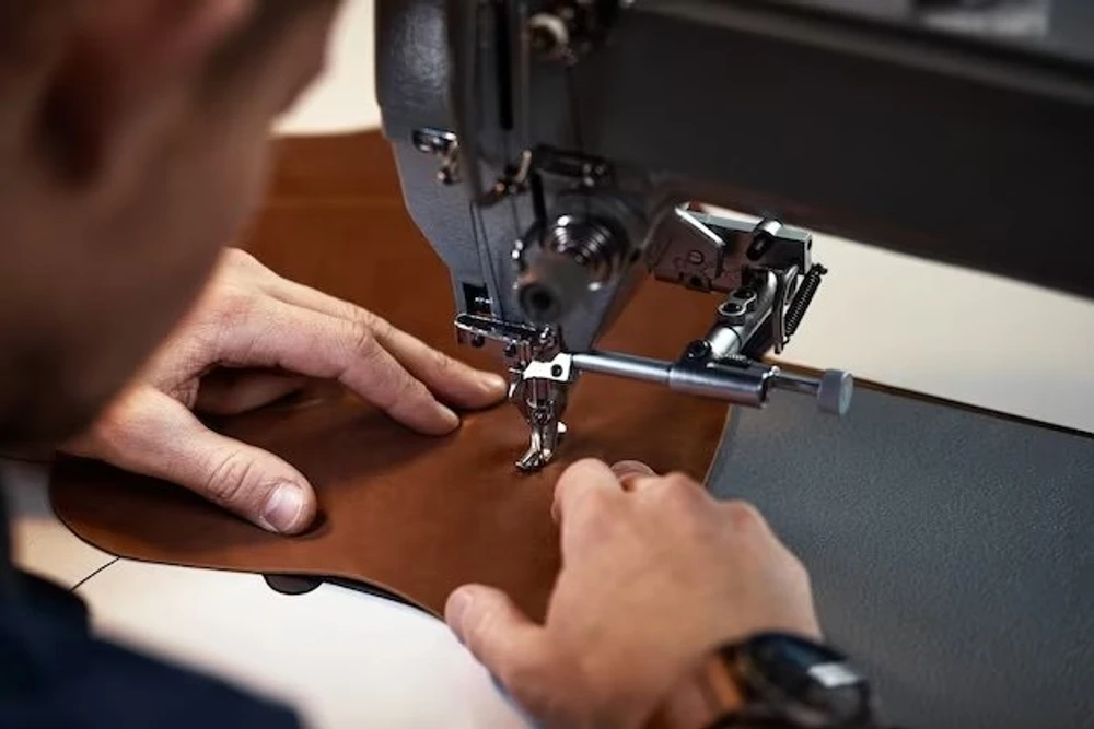 A machinist guiding a brown leather panel through a heavy industrial sewing machine