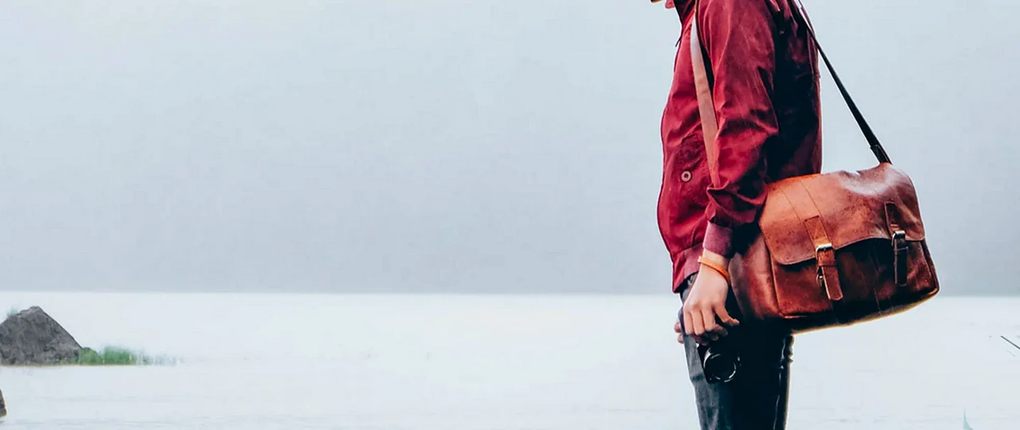 A person in a red jacket crossing an open shore with a brown leather flap messenger bag on the shoulder