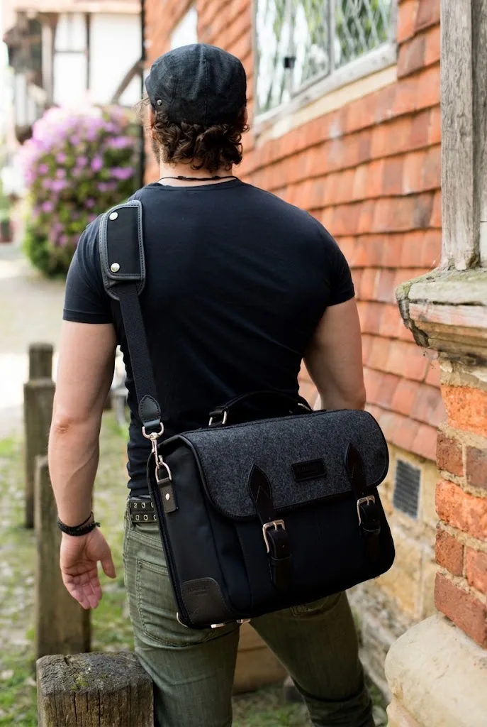 A black wool-flap messenger bag worn across the back of a man in a cap, on a cobbled lane beside a brick wall