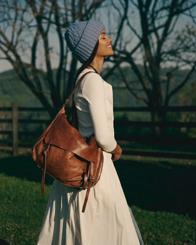 A tan leather messenger bag on the shoulder of a woman in a knit hat, crossing open ground beneath bare trees