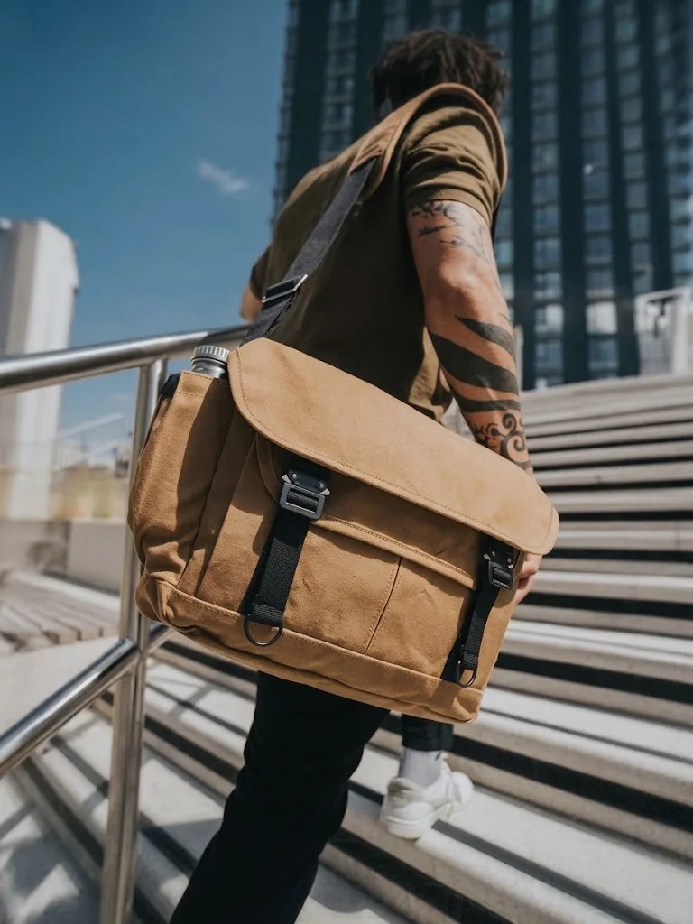 A tan canvas messenger bag slung low at the hip of a man crossing a stepped plaza in the sun
