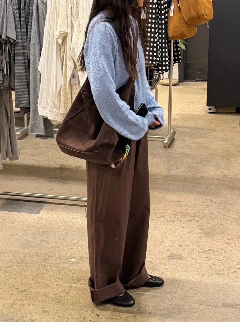 A brown suede shoulder bag carried under the arm of a woman standing among clothing rails in a store