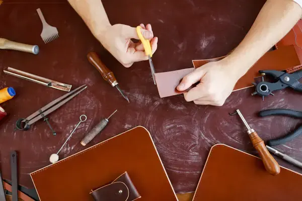 Hands cutting a leather panel with a craft knife on a bench surrounded by stitching awls, dividers and hole punches