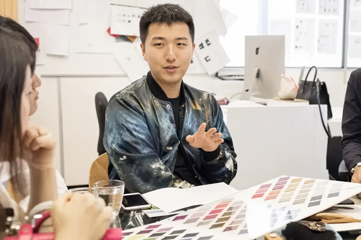 A factory contact talking across a table of open colour cards to a buyer seated opposite, sample sheets pinned on the wall behind