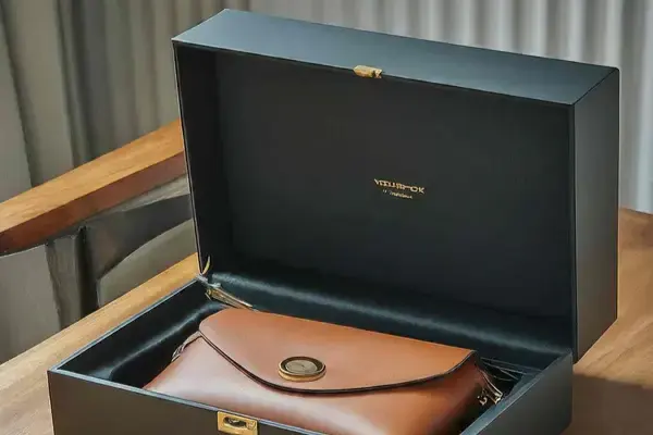 A leather handbag resting in an open black presentation box with a coral lining, on a wooden bench