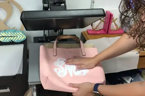 A heat press transferring a white monogram onto the front of a pink canvas tote