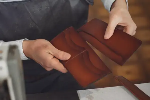 A leatherworker holding two folded panels of tanned hide up to the light beside a stack of swatches
