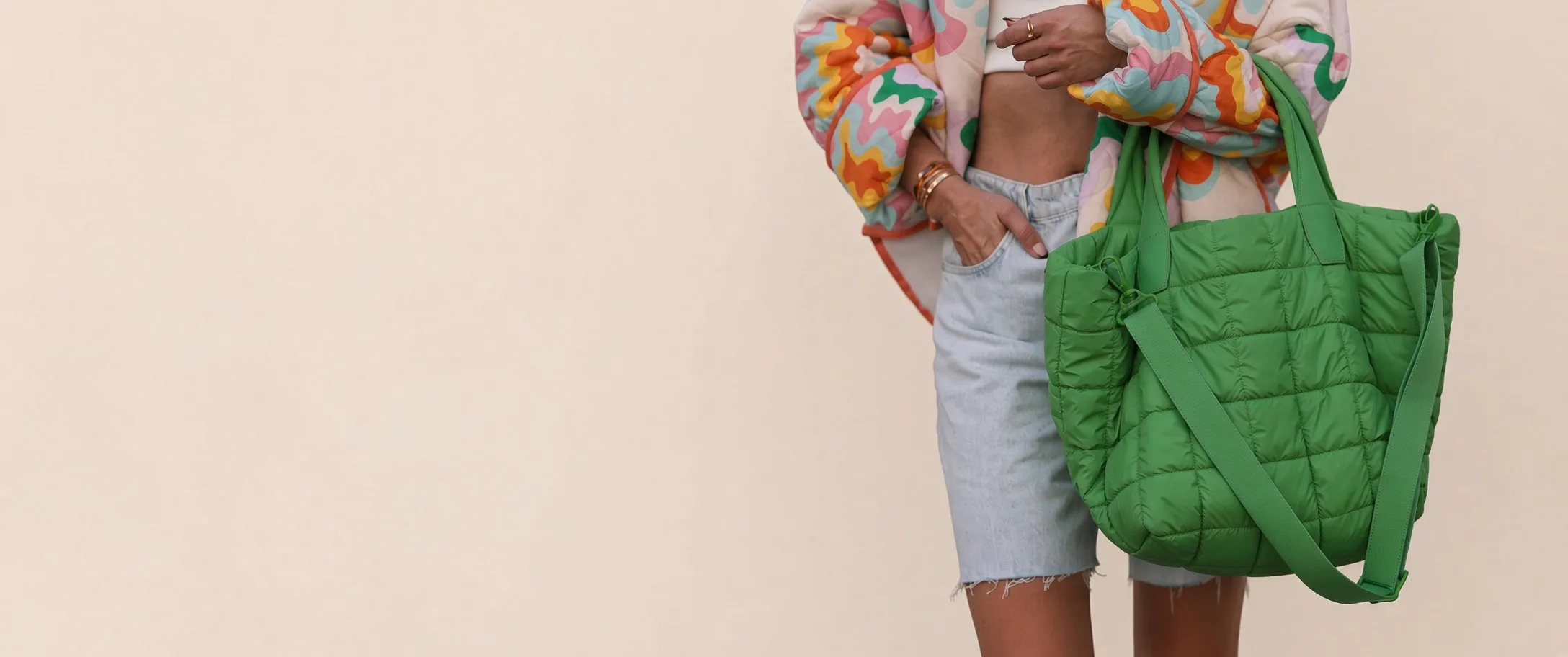 A model in a patterned jacket holding a green quilted tote with a crossbody strap, against a pale studio wall