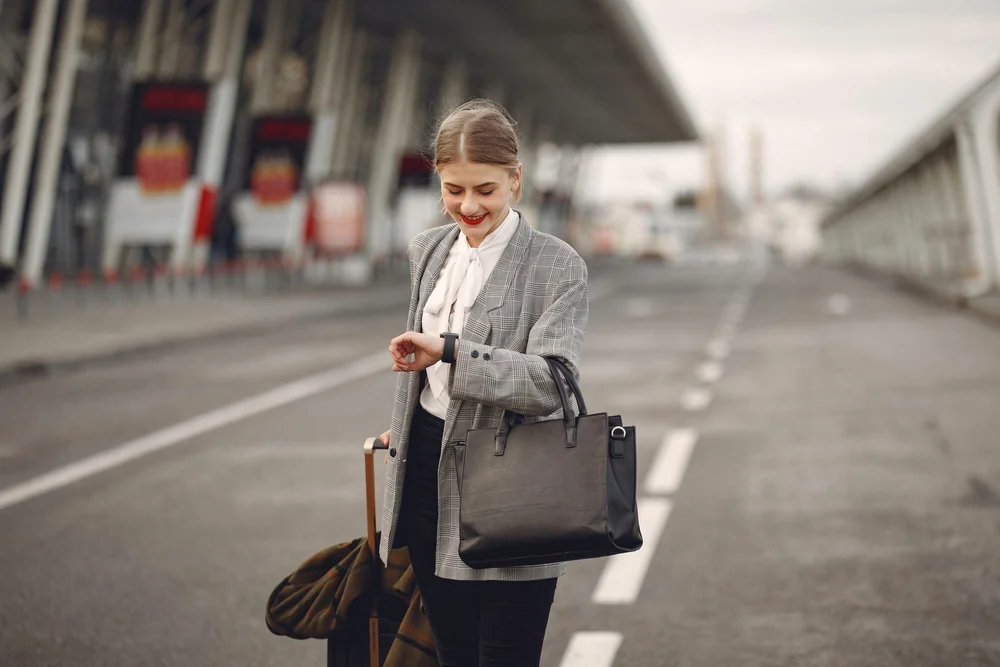 A traveller in a checked blazer checking her watch on an empty airport drop-off road, a black tote on one shoulder and a wheeled case beside her