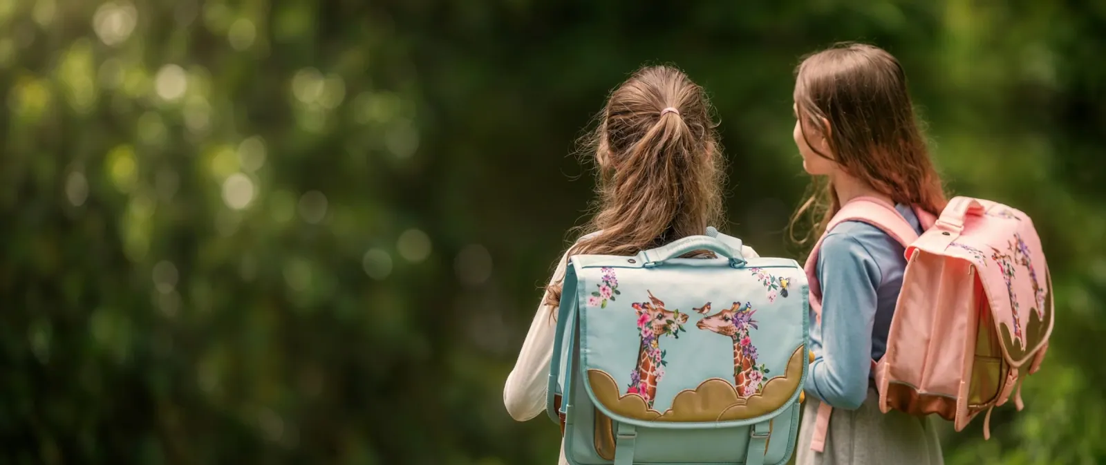 Two school students seen from behind on a tree-lined path, wearing differently sized school backpacks with a top handle and a printed front panel