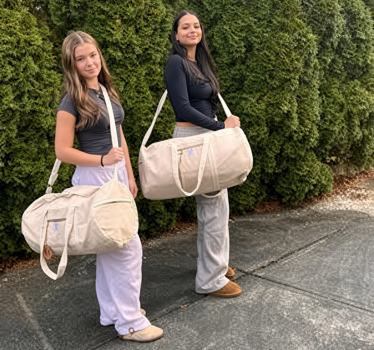 Two women walking a paved path, both carrying a cream travel duffle