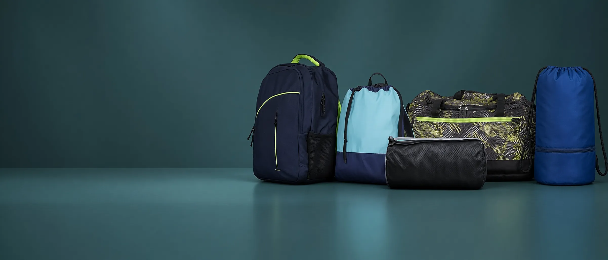 Five sports bags in a row against a deep teal studio wall — a navy backpack, a pale-blue drawstring pack, a black barrel duffle, a patterned holdall and a blue stuff sack