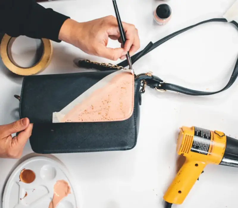 A craftsperson edge-coating a black leather handbag with a brush beside a heat gun and a painter palette