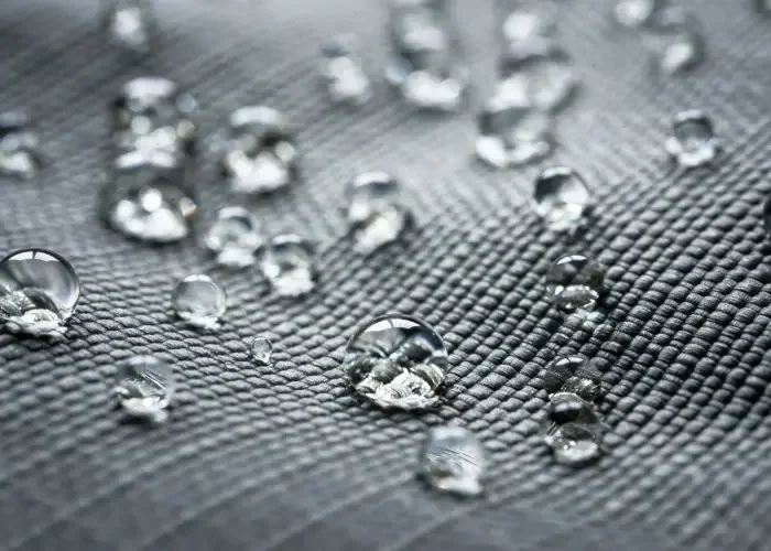Water beading on a tightly woven grey coated shell fabric at close range, the droplets standing proud of the weave
