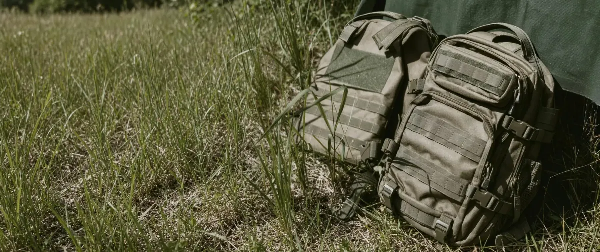 Two olive tactical backpacks resting against a green sheet in long grass, the front pack showing its MOLLE webbing panels, side compression buckles and twin zip pulls
