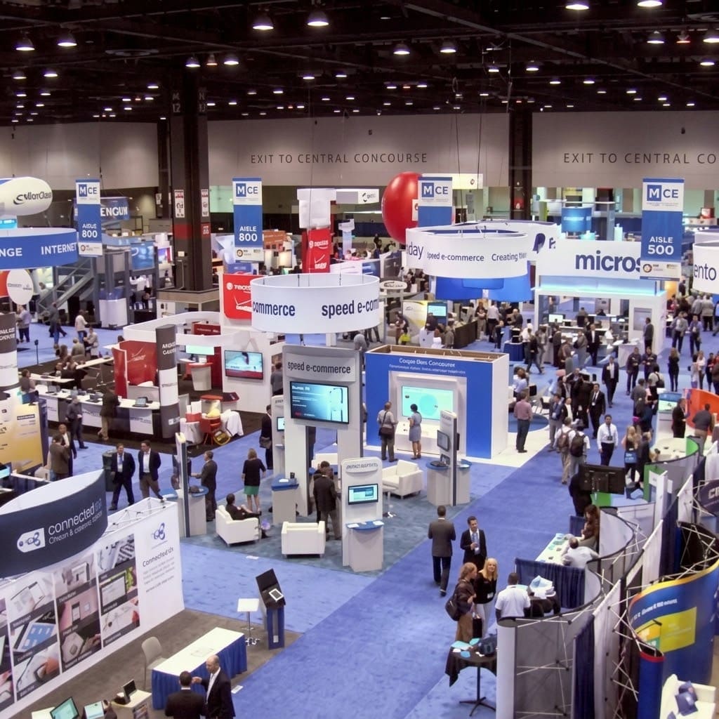 A crowded exhibition hall seen from above, ranked rows of branded booths along a blue aisle carpet