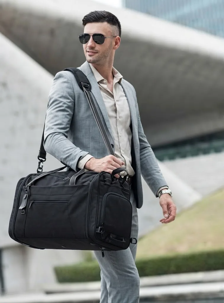 A man in a grey suit crossing a plaza with a black duffel travel bag over his shoulder